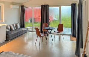 Ein modernes Wohnzimmer mit einem grauen Sofa, zwei Sesseln, einem kleinen Tisch und einer großen Schiebeglastür, die zu einem Garten führt.