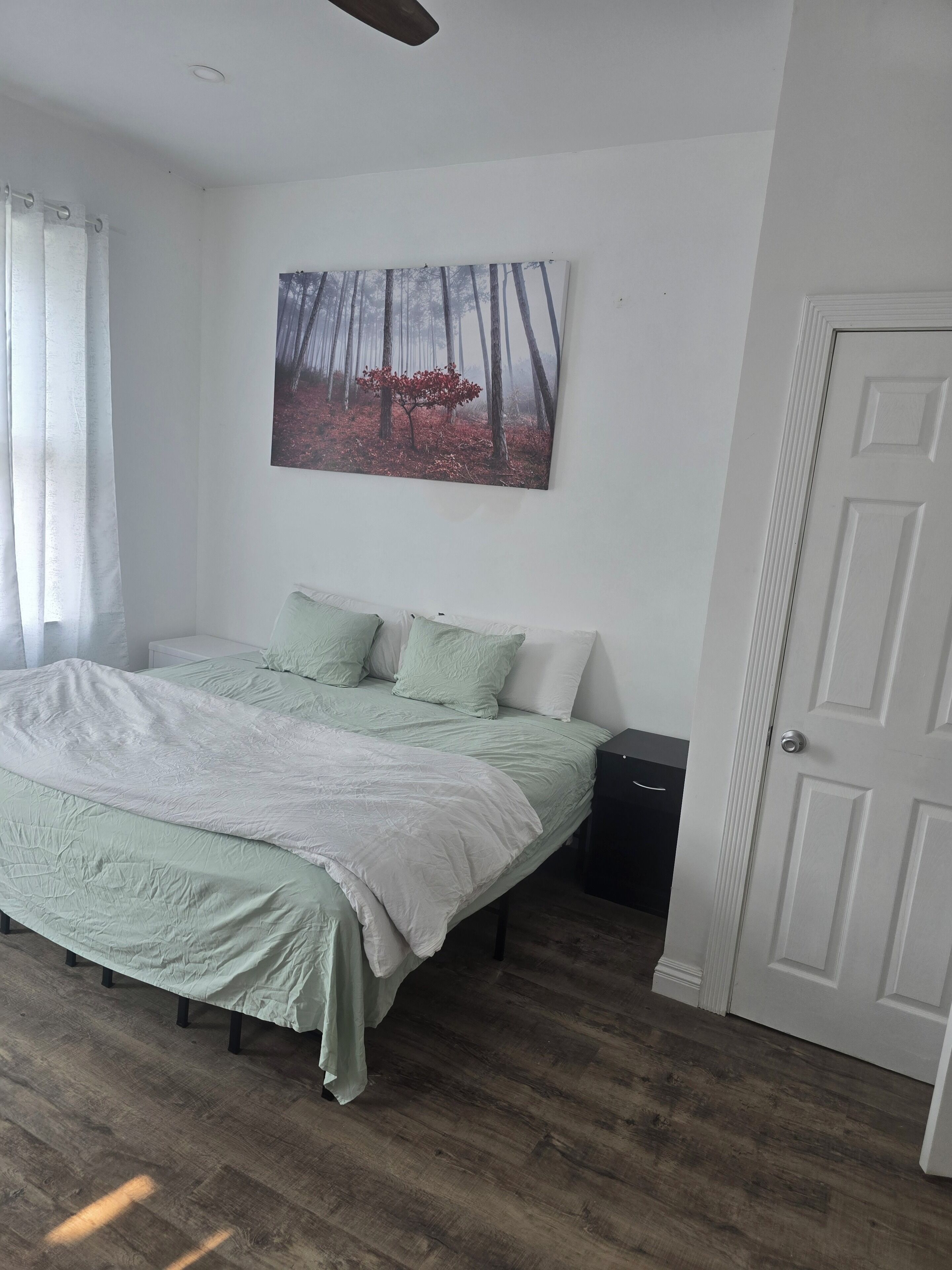 Une chambre à coucher avec un lit, un tableau, une porte et une fenêtre avec des rideaux.