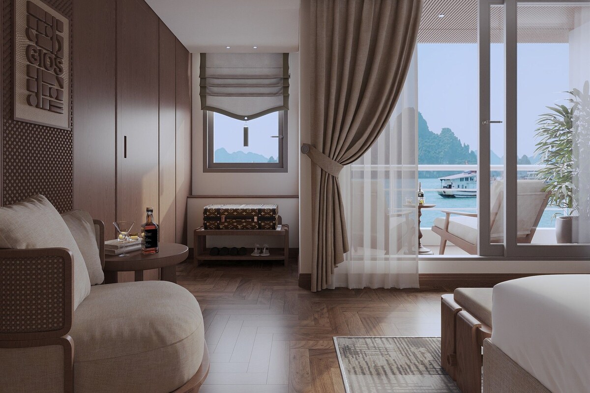 A modern hotel room with a large window, a sofa, a bed, a small table with a bottle, and a suitcase.