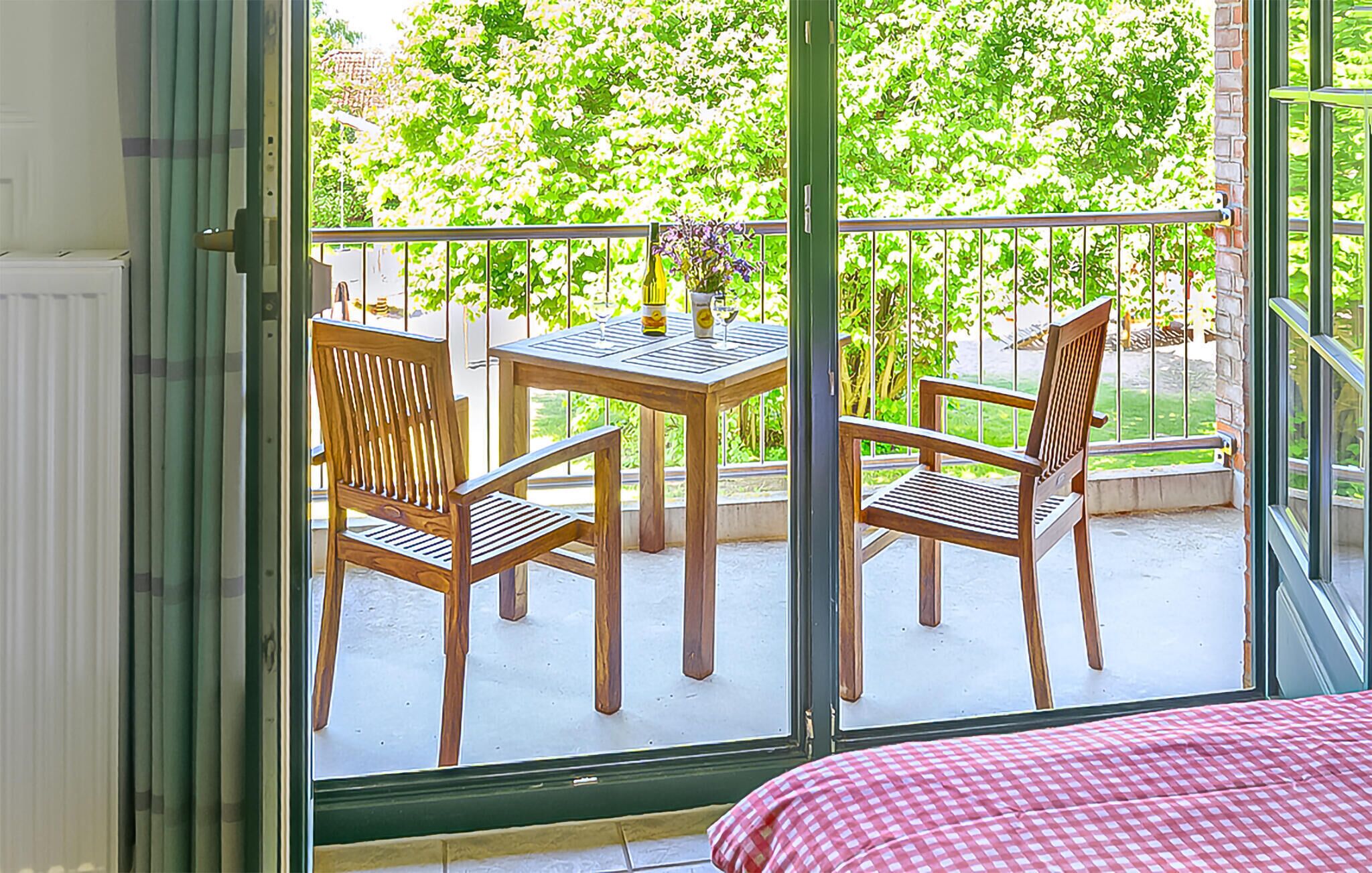 Un balcon avec deux chaises en bois et une table, une nappe à carreaux et une vue sur la verdure, visible à travers une porte vitrée.