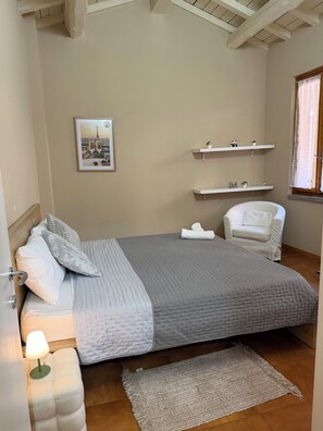 A bedroom with a bed, a chair, a bedside table with a lamp, a picture on the wall, and a window with a curtain. - Dragonfly Pavia (Pavia)