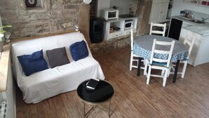 Een knus vertrek met een witte bank, een kleine ronde tafel, een eettafel met stoelen, een keuken met witte kasten en een stenen muur.