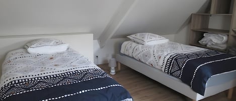 A bedroom with two beds, each with patterned bedding and pillows, a bedside table with a lamp, and a wooden shelf with folded clothes.