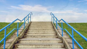 Unos escalones de hormigón con barandillas azules que suben por una colina cubierta de césped bajo un cielo despejado.