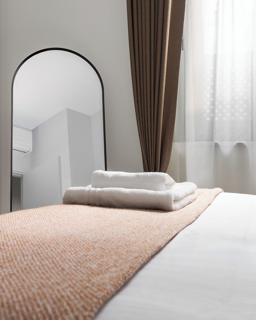 A neatly made bed with a white duvet and a folded white towel, a brown textured blanket, a black-framed mirror, and a window with sheer curtains.