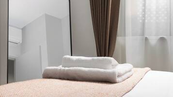 Una cama bien hecha con un edredón blanco y una toalla blanca doblada, una manta marrón con textura, un espejo con marco negro y una ventana con cortinas traslúcidas.