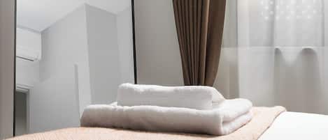 Una cama bien hecha con un edredón blanco y una toalla blanca doblada, una manta marrón con textura, un espejo con marco negro y una ventana con cortinas traslúcidas.