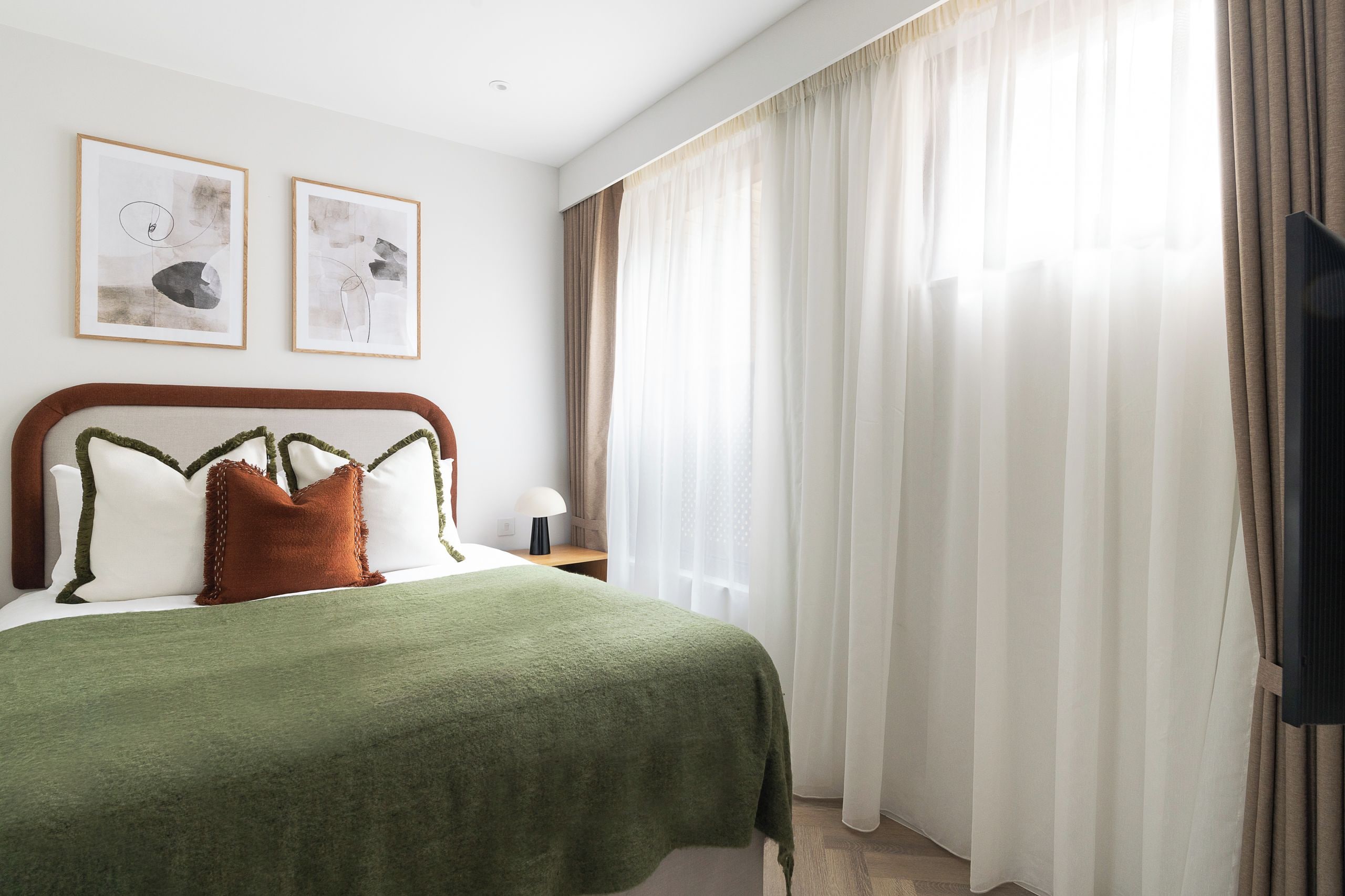 Ein Hotelzimmer mit einem Bett, einem Nachttisch, einer Lampe und zwei gerahmten Kunstwerken an der Wand.