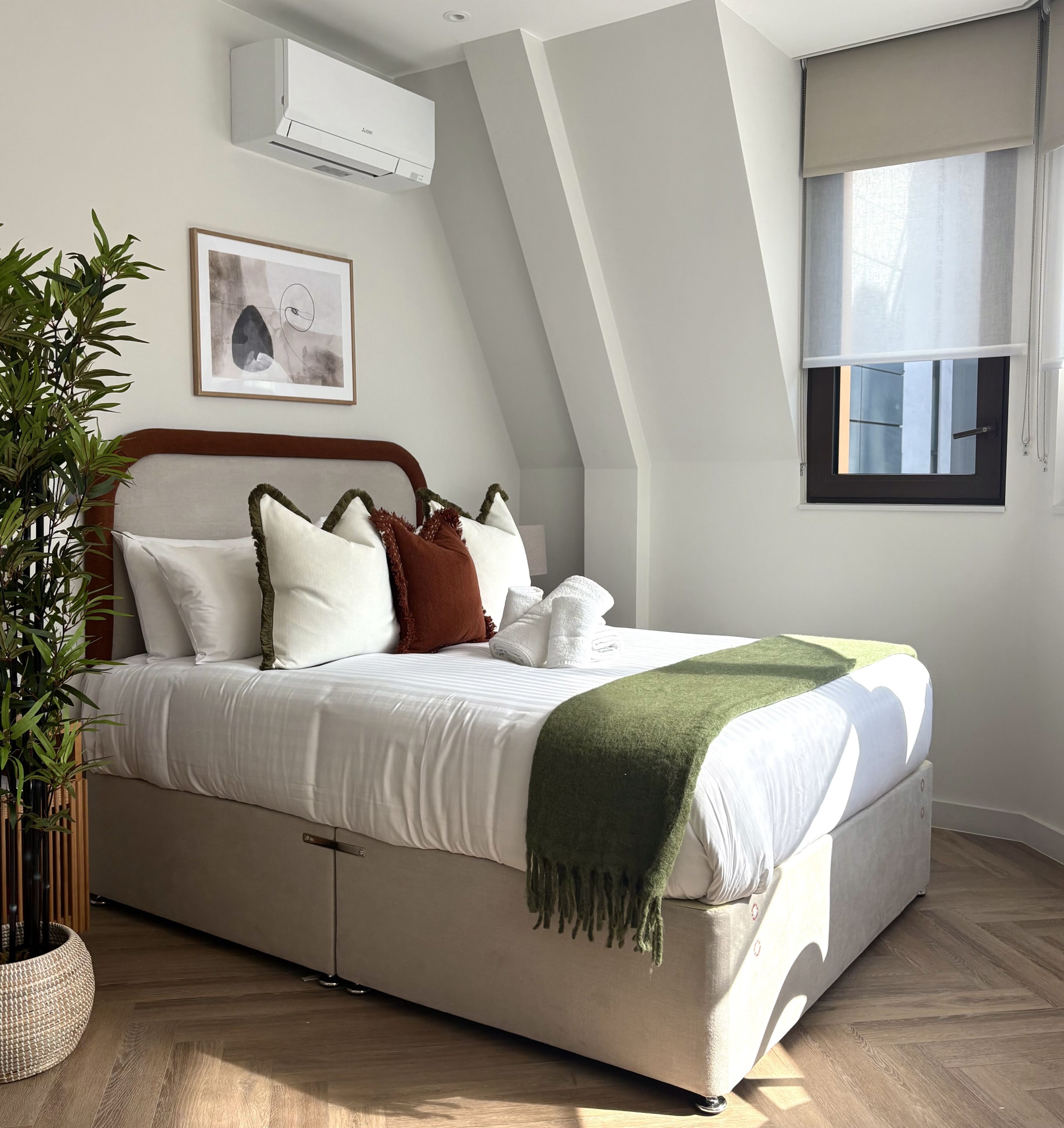 A bedroom with a bed, pillows, a blanket, a plant, a framed picture, and a window.