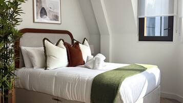 A bedroom with a bed, pillows, a blanket, a plant, a framed picture, and a window.
