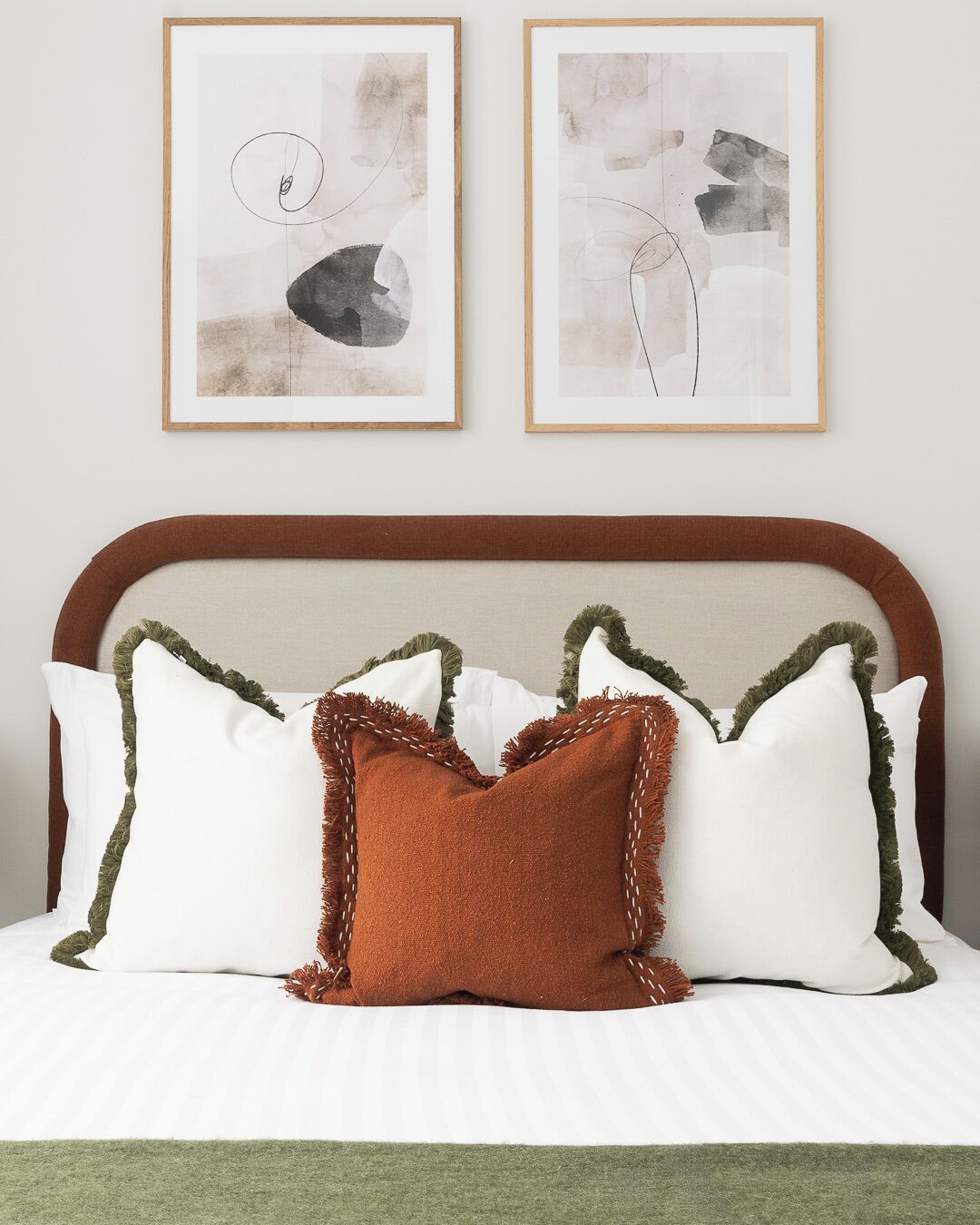 A bed with white bedding, a brown accent pillow, and green trim on the headboard. Two framed abstract artworks are hung above the bed.