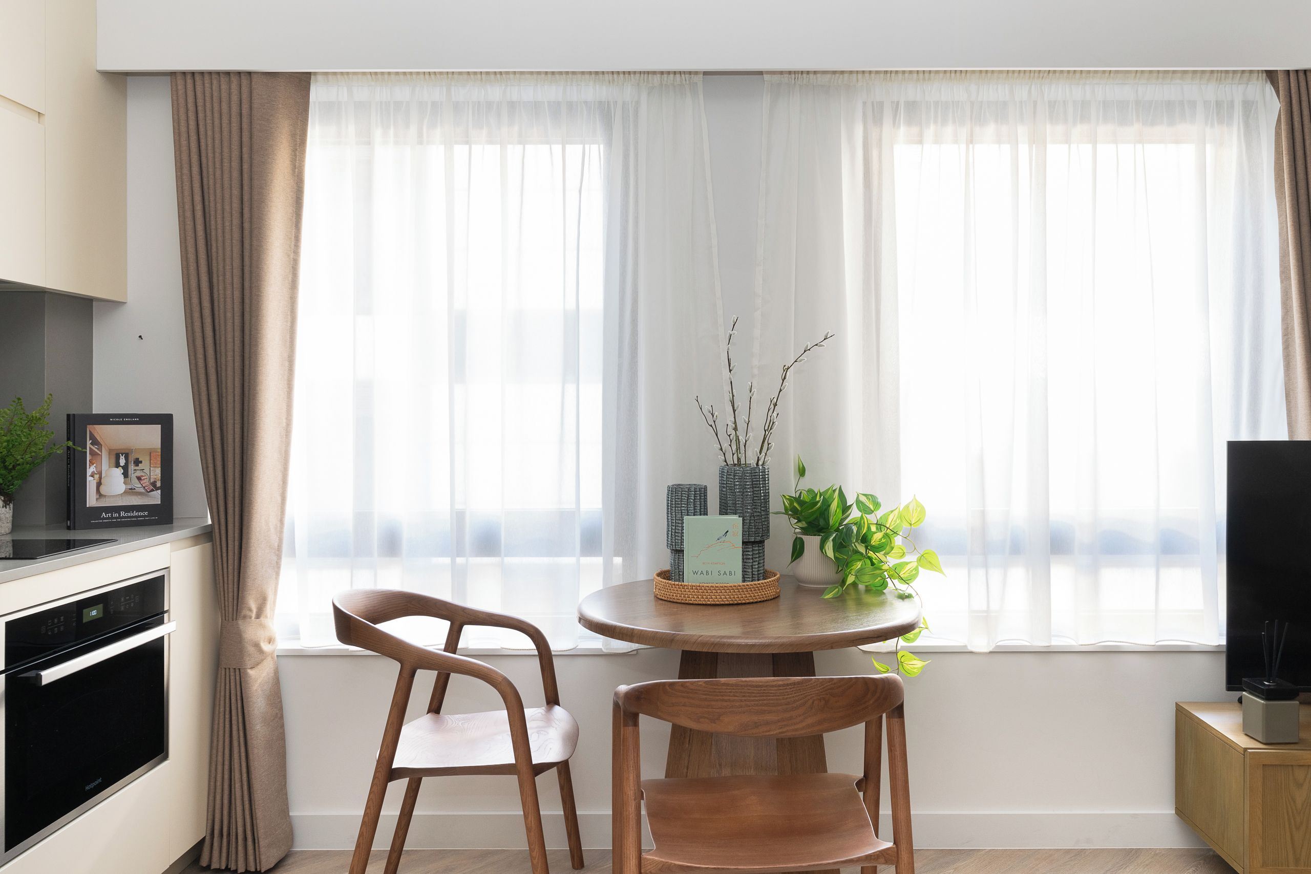 A modern kitchen with a built-in oven, a dining area with two wooden chairs, a round table, and a window with sheer curtains.