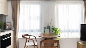 A modern kitchen with a built-in oven, a dining area with two wooden chairs, a round table, and a window with sheer curtains.