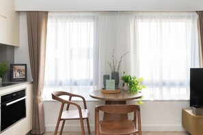 Una cocina moderna con horno integrado, una zona de comedor con dos sillas de madera, una mesa redonda y una ventana con cortinas ligeras.