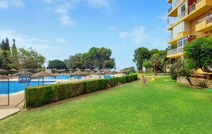 Zona de piscina con sombrillas, césped cuidado y un edificio con balcones.