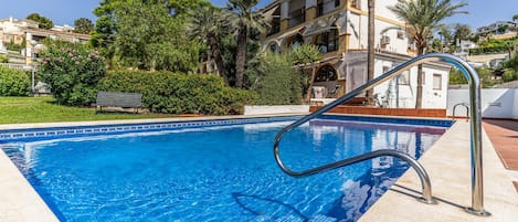 A swimming pool with a metal ladder, surrounded by greenery and a building with balconies.