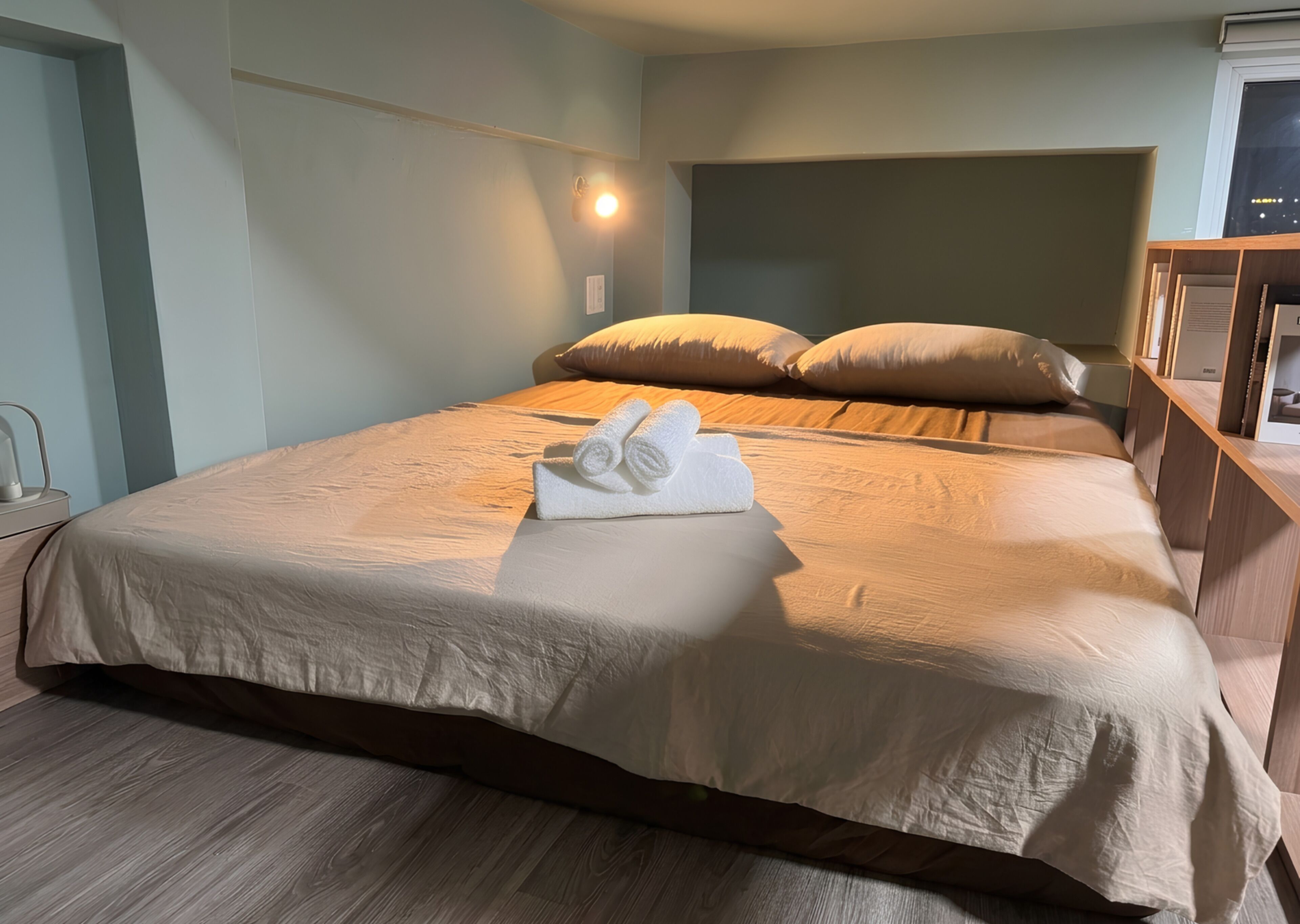 Ein ordentlich bezogenes Bett mit einem gefalteten Handtuch, einem Nachttisch und einer Wandleuchte.