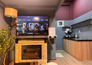 A modern living room with a fireplace, a flat-screen TV mounted on the wall, a kitchen area with a sink and countertop, and a decorative plant. - Àla Homestay Binh Thanh - Loft Studio with PS5 (Ho Chi Minh City)