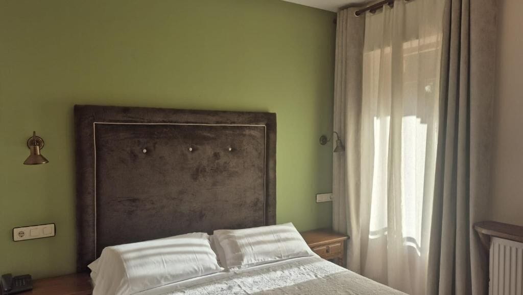 A room with a bed, a nightstand, a wall-mounted light, and a window with curtains.