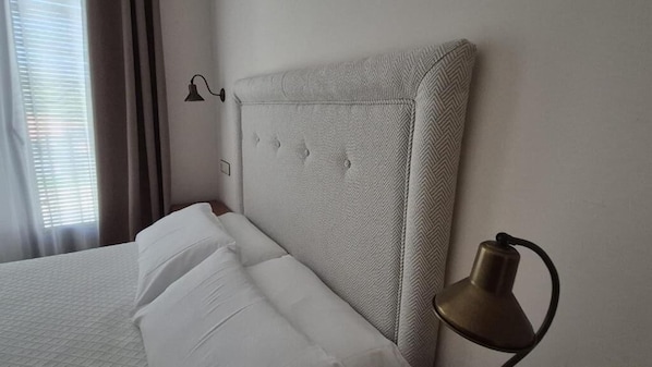A hotel room with a bed, a headboard, a wall-mounted lamp, and a window with curtains.
