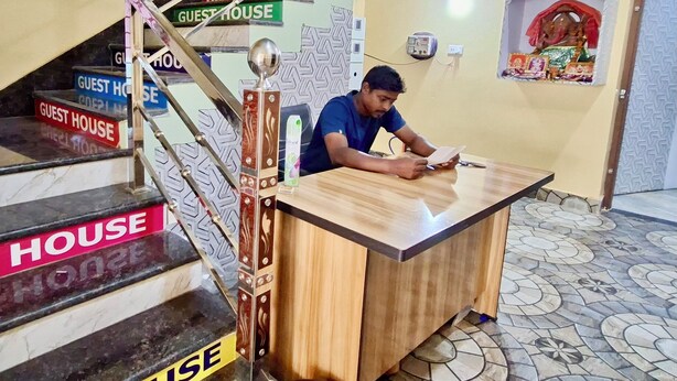 Un hombre sentado en un escritorio de madera en una habitación con piso de baldosas y una escalera con letreros que dicen 'GUEST HOUSE'.