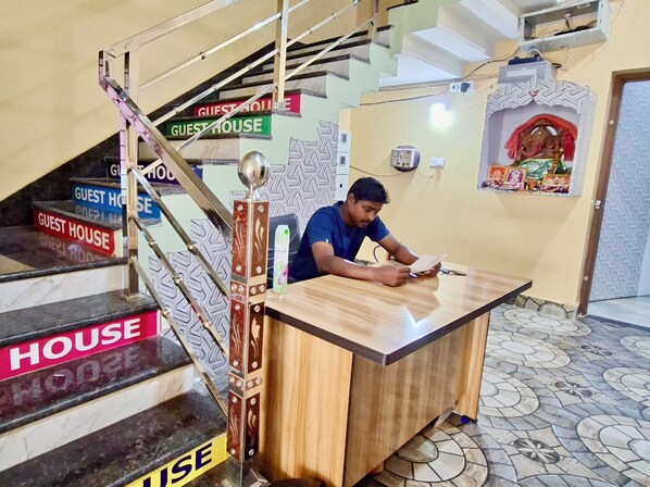 A man seated at a wooden desk in a room with a tiled floor and a staircase with 'GUEST HOUSE' signs.