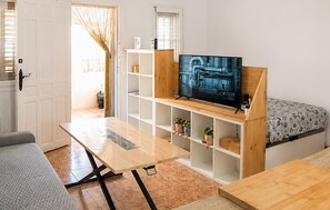Een ruimtelijk ingerichte leefruimte met een televisie op een houten dressoir, een bank, een klein tafeltje en een raam met zonwering.
