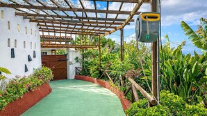 Ein überdachter Außenbereich mit einem Grünstreifen, umgeben von üppiger Vegetation und einem weißen Gebäude mit einer Holzpergola.