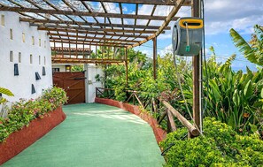 Ein überdachter Außenbereich mit einem Grünstreifen, umgeben von üppiger Vegetation und einem weißen Gebäude mit einer Holzpergola.