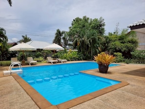 Una piscina rectangular con superficie de agua azul cristalina, rodeada por una solana con baldosas. Hay sillones de playa y sombrillas, y una planta en maceta cerca de la piscina.