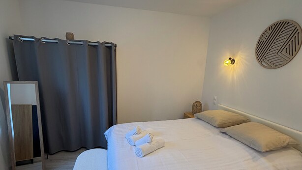 A neatly made bed with pillows and a folded towel, a bedside table, a wall-mounted light fixture, and a decorative wall piece. - 4★ Cozy Apartment in Contrexéville Thermal Baths (Contrexéville)