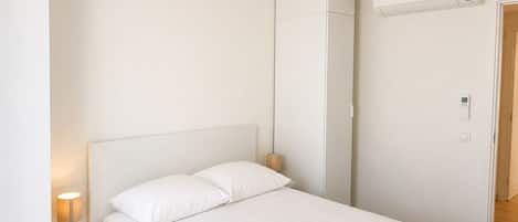 Een minimalistische slaapkamer met een bed, bedlampje, kledingkast en een airconditioner.