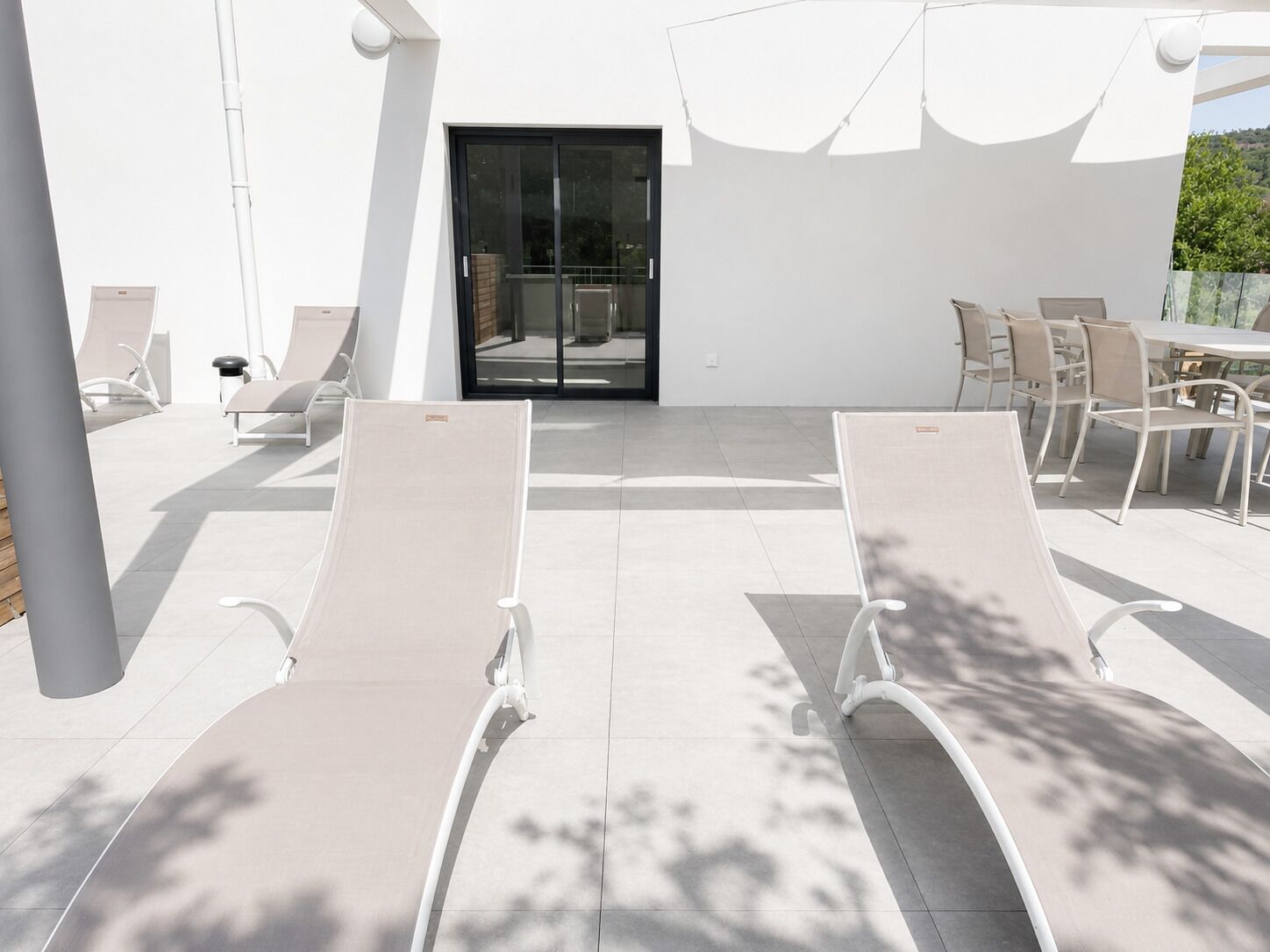 Un espacio al aire libre iluminado por el sol con sillones de descanso y una mesa, una entrada por puertas de vidrio y fachada de un edificio blanco.