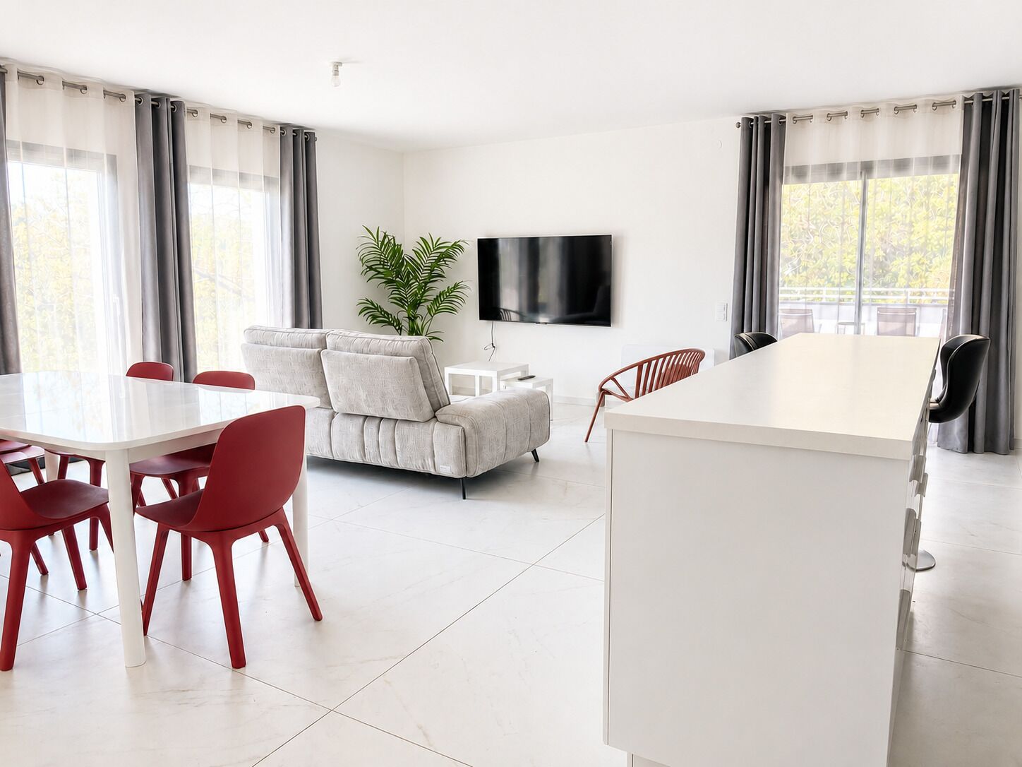 Un salon moderne comprenant un canapé, une table à manger avec des chaises, un téléviseur à écran plat fixé au mur et une grande fenêtre avec des rideaux.