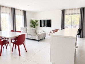 A modern living room with a sofa, a dining table with chairs, a flat-screen TV mounted on the wall, and a large window with curtains. - 3-bedroom apartment measuring 86 m², with a 70 m² rooftop terrace and a private pool (Saint-Raphaël)