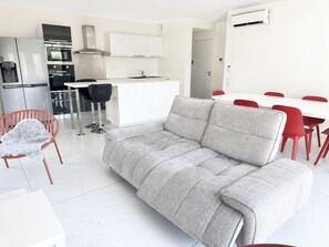 A modern living room with a grey sofa, a dining area with red chairs, a kitchen with white cabinets, and a built-in oven. - 3-bedroom apartment measuring 86 m², with a 70 m² rooftop terrace and a private pool (Saint-Raphaël)