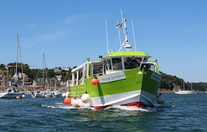 A green boat with the name 'LE PASSEUR DU TRIEUX' on its side, moving through a harbor with other boats and buildings in the background. - Nice apartment in Lezardrieux with house sea view (Lezardrieux)