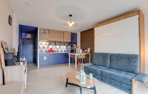 A modern living room with a blue sofa, a wooden coffee table, and a kitchen area with blue cabinets and a hanging light fixture. - Nice apartment in Lezardrieux with house sea view (Lezardrieux)