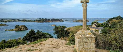 A stone cross on a hill overlooking a coastal area with a body of water and distant land.