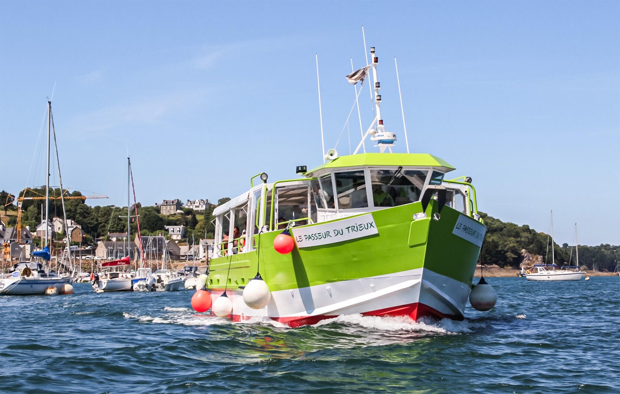 Un barco blanco y verde llamado 'Le Passeur du Trieux' en el agua, con otros barcos y casas al fondo.