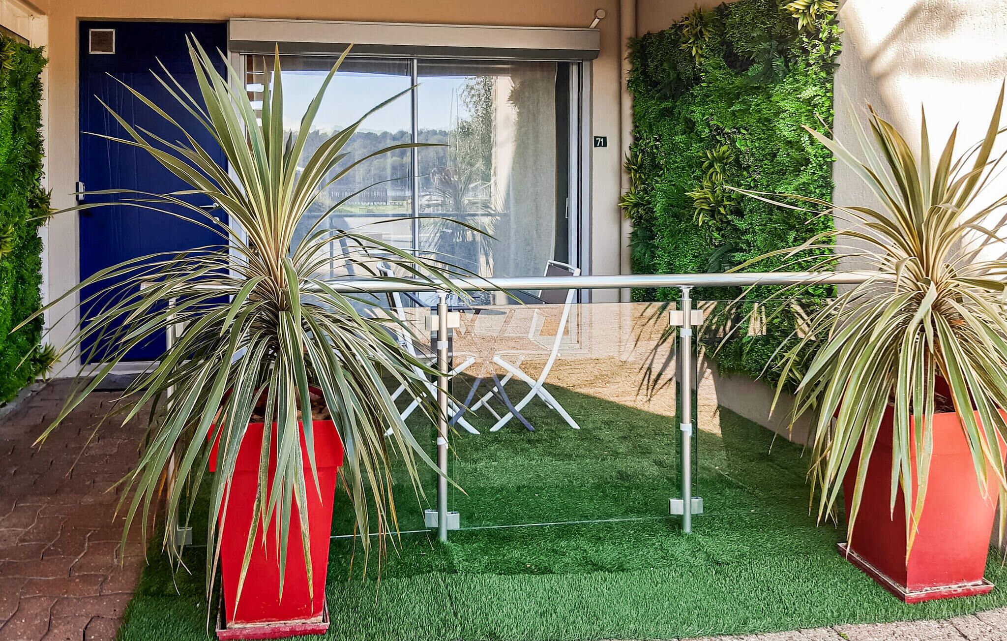 Un espacio exterior moderno con césped artificial, dos plantas grandes en macetas, una barandilla metálica y una puerta azul.