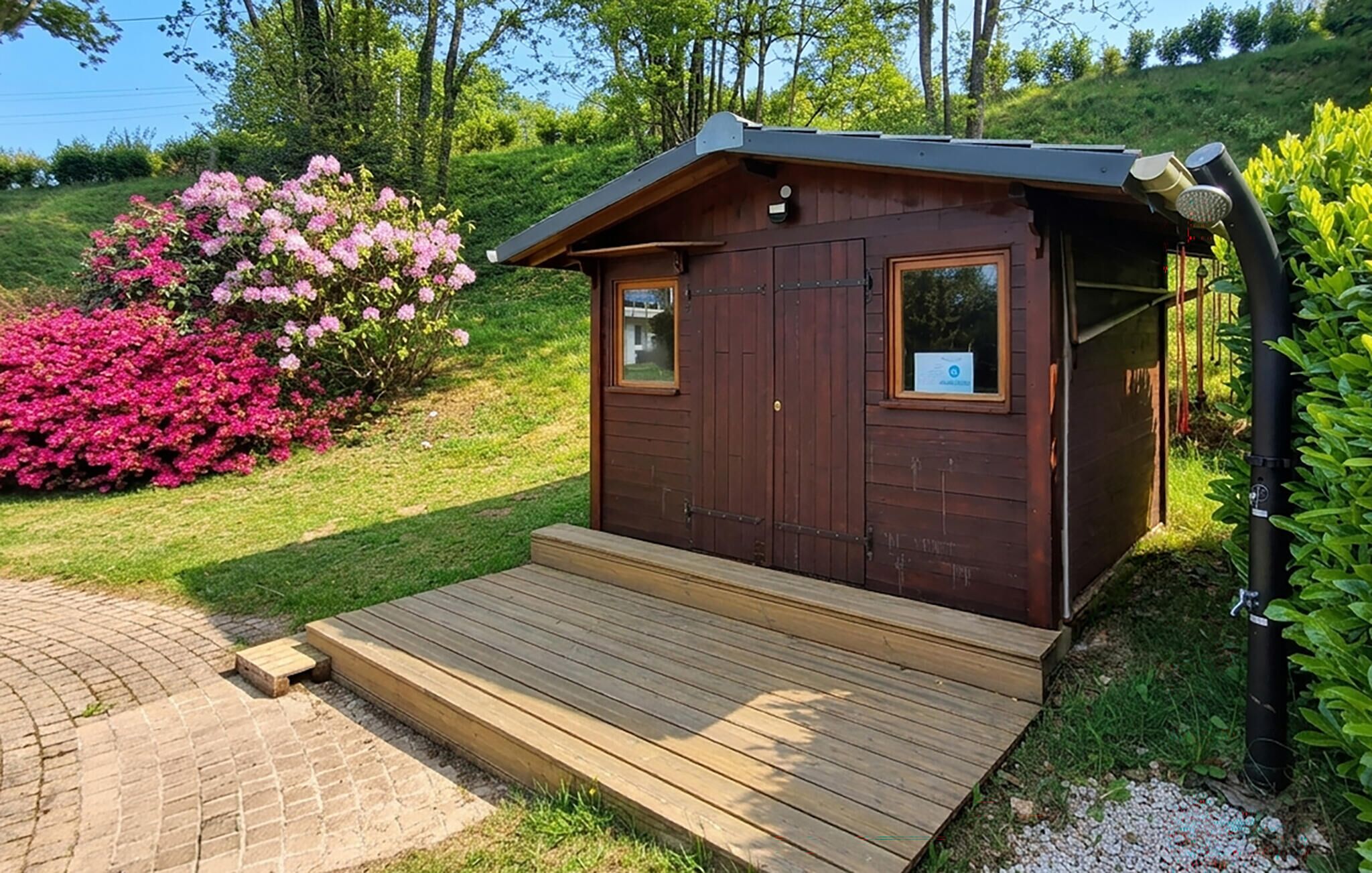 Una cabaña de madera con un porche pequeño, rodeada de abundante follaje y arbustos florecientes.