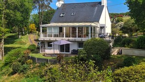 Une maison à deux étages dotée d’un balcon, entourée d’une végétation luxuriante et sous un ciel dégagé.