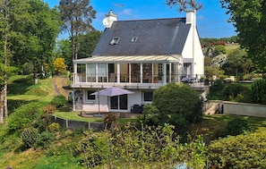 Ein zweigeschossiges Haus mit Balkon, umgeben von üppiger Vegetation und blauem Himmel.