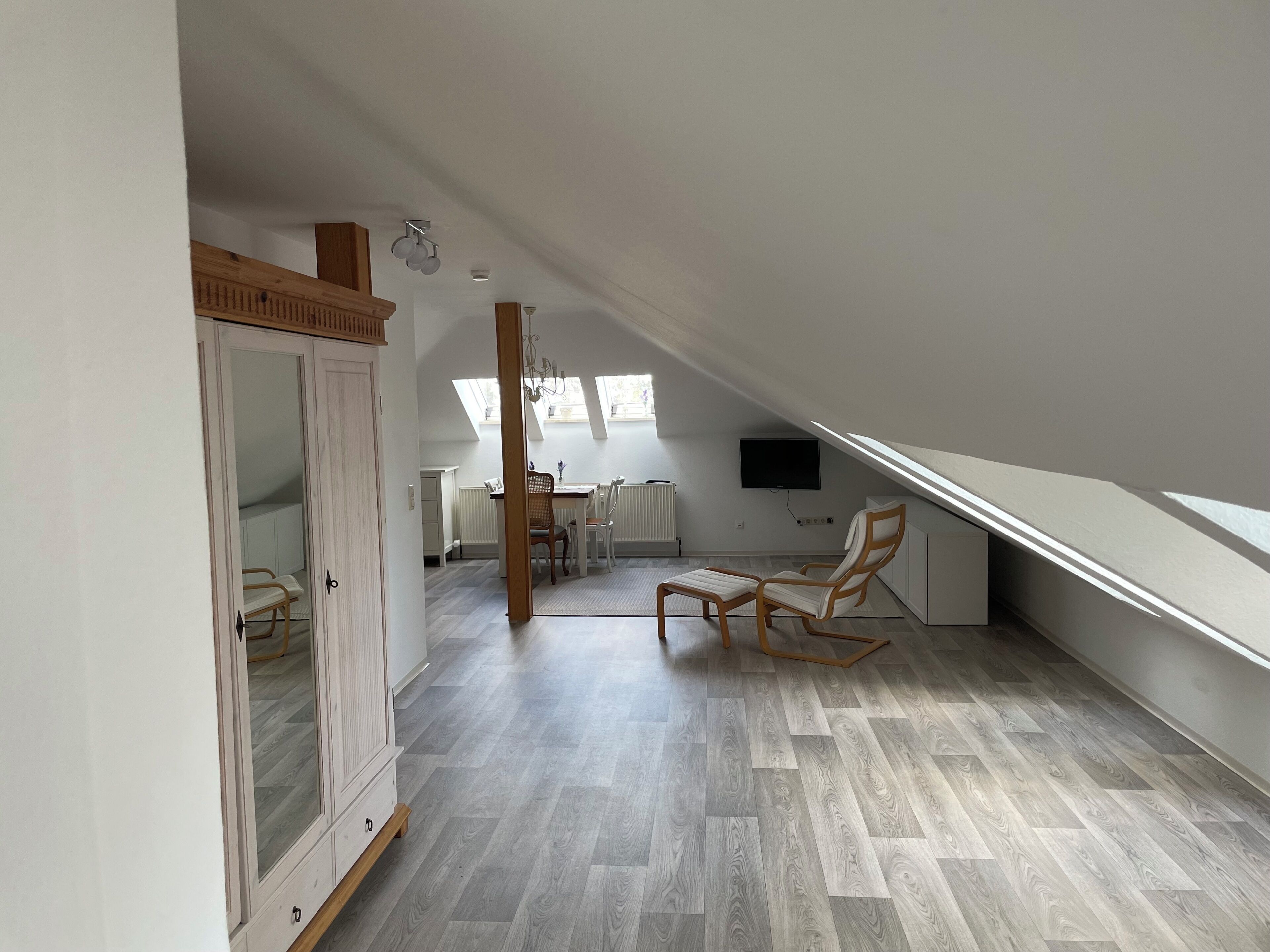 Amplia habitación en el ático con techo alto, piso de madera, un gran armario, zona de comedor con mesa y sillas, y televisor montado en la pared.