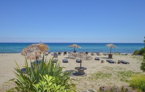 Ein Strand mit Liegestühlen und Reetdach-Sonnenschirmen, feiner Sand und ein klarer, blauer Himmel.