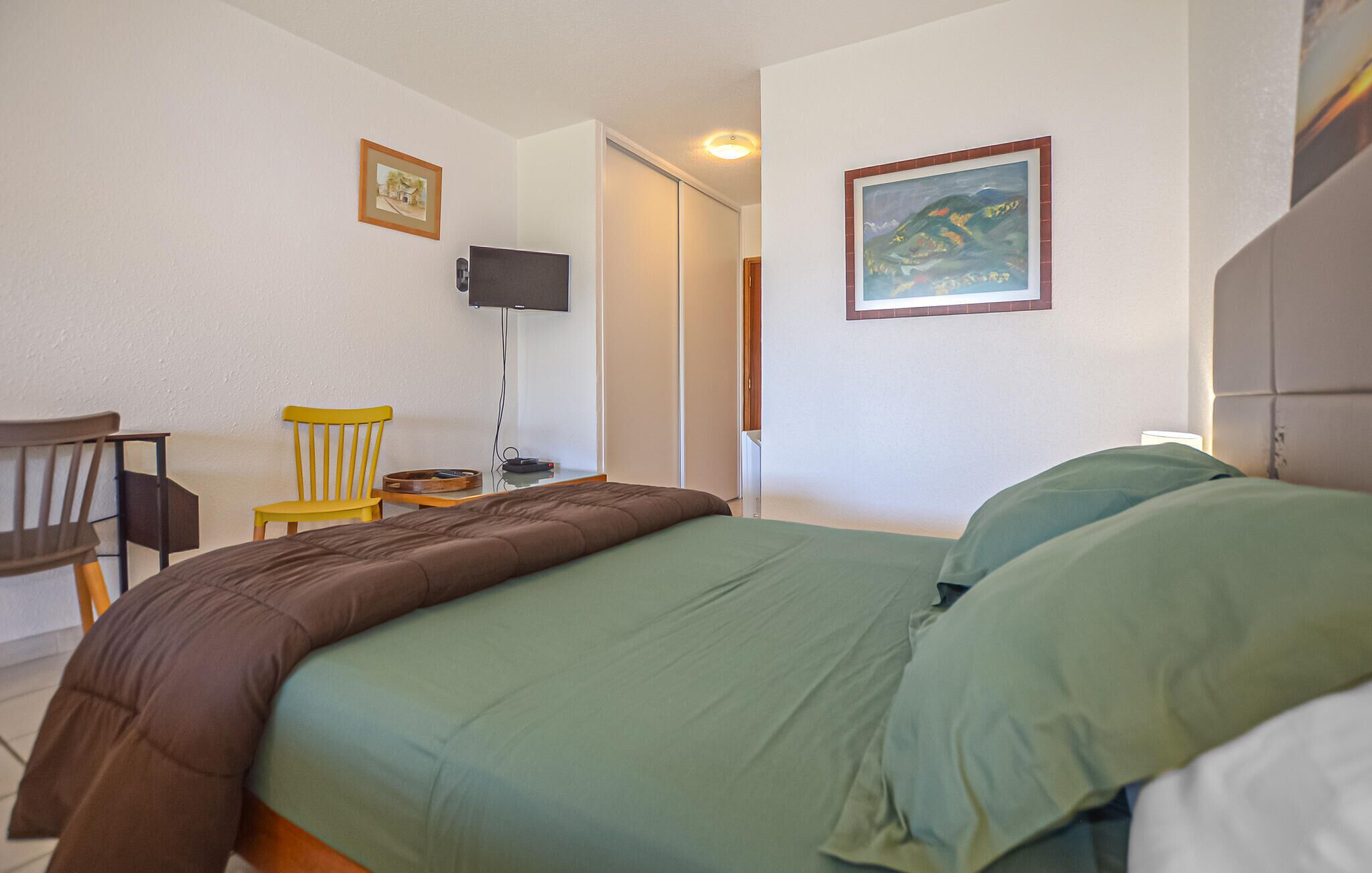 Une chambre à coucher comprenant un lit, une chaise, un bureau, un téléviseur et deux tableaux encadrés accrochés aux murs.
