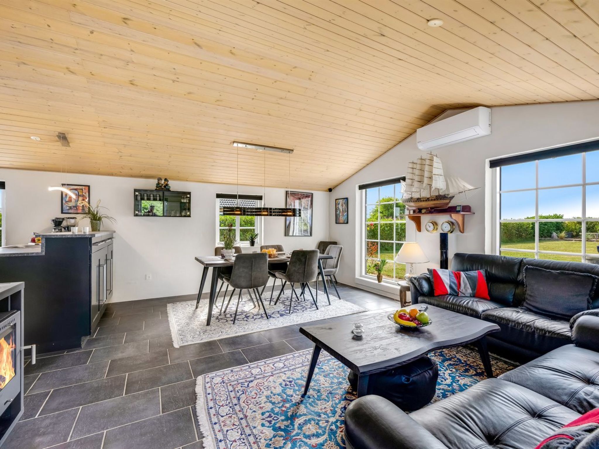 A spacious living area with a fireplace, a dining table with chairs, a black leather sofa, and a large window offering a view of the outdoors.