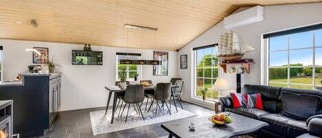 A spacious living area with a fireplace, a dining table with chairs, a black leather sofa, and a large window offering a view of the outdoors.