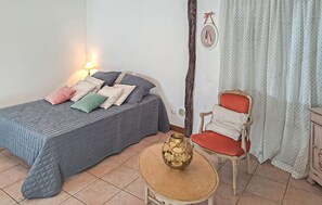 Ein Schlafzimmer mit Bett, Kissen, einem Sessel, einem kleinen Tisch, einer Lampe und Vorhängen.